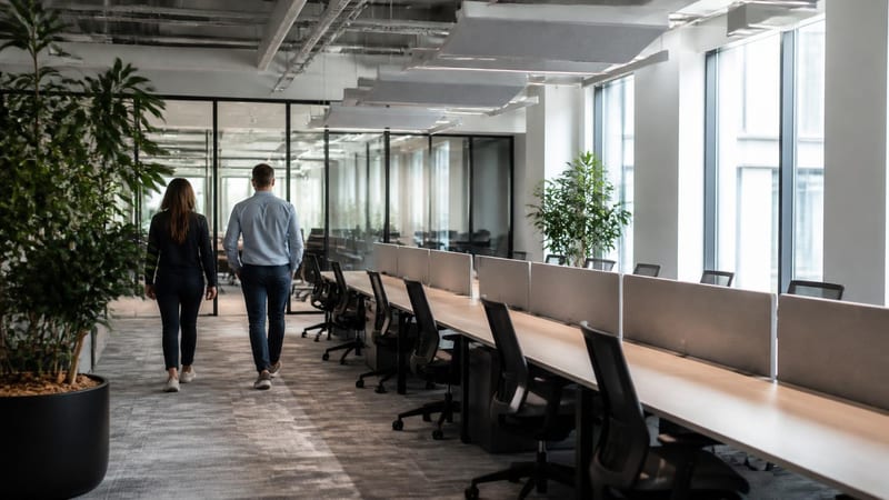 A commercial office floor of long shared desks, two people walking through at a distance